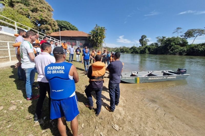 Pessoas reunidas na margem de um rio ao lado de um barco de alumínio com a inscrição Marinha do Brasil.