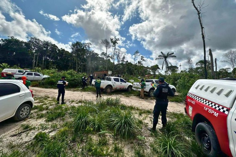 Foto ao ar livre com viaturas policiais, agentes de segurança fardados e civis em uma estrada de terra cercada por vegetação e árvores, sob um céu com nuvens brancas.