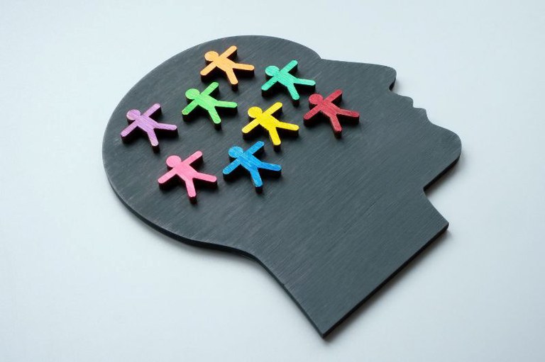 Arte de uma silhueta de perfil de uma cabeça humana, de cor cinza escura, sobre um fundo branco. No interior da silhueta, na área do cérebro, estão espalhadas diversas figuras pequenas e coloridas em formato de pessoas.