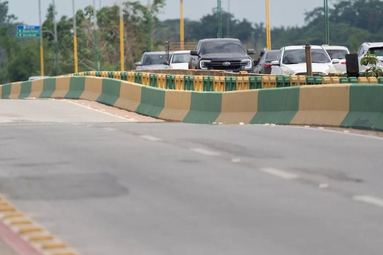 Uma visão em nível da estrada de uma ponte com barreiras amarelas e verdes, onde veículos aguardam em engarrafamento no fundo.