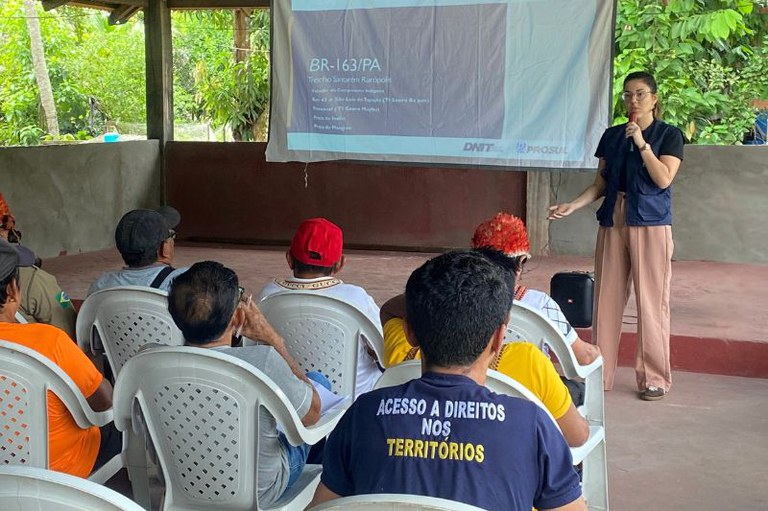 Foto da procuradora da República Thaís Medeiros da Costa ao microfone diante de uma tela que exibe "BR-163/PA". Na plateia, várias pessoas estão sentadas em cadeiras brancas, vestindo camisetas coloridas, incluindo uma com a frase "Acesso a direitos nos territórios". Também há pessoa indígena com cocar de penas vermelhas. O ambiente é um espaço coberto ao ar livre, cercado por vegetação.
