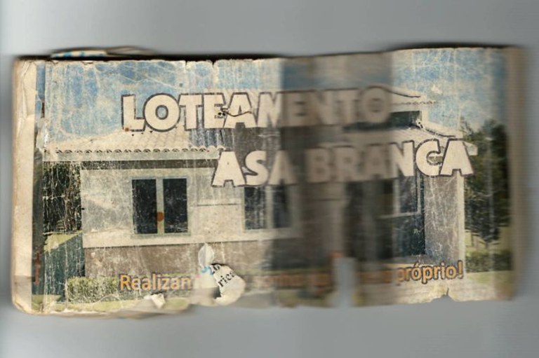 Fotocópia de boleto de papel antigo, dobrado e desgastado. O anúncio é do "Loteamento Asa Branca" e mostra a foto de uma casa. As bordas do papel estão rasgadas e a imagem está envelhecida.