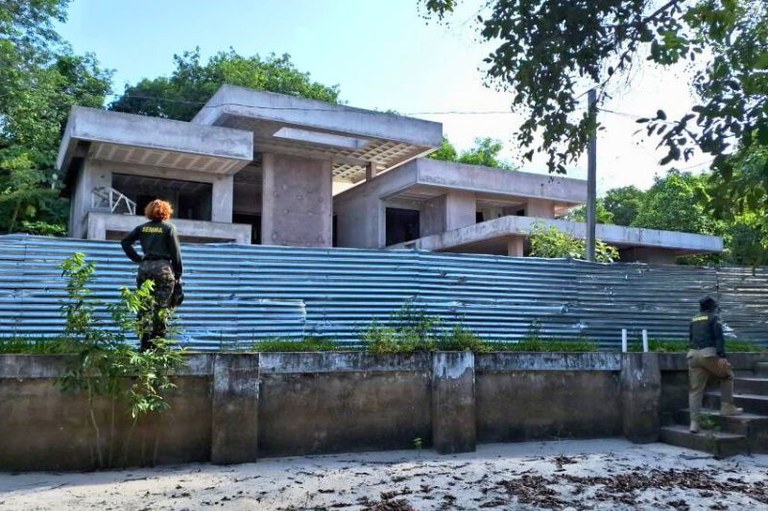 Agentes da Semma fiscalizam obra de casa moderna em concreto bruto, cercada por tapume metálico e vegetação em Alter do Chão.