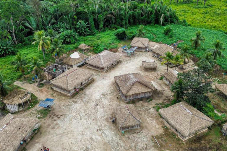 Uma fotografia aérea de uma pequena aldeia indígena com várias cabanas de telhado de palha, situada em uma clareira de terra cercada por uma densa floresta verde.