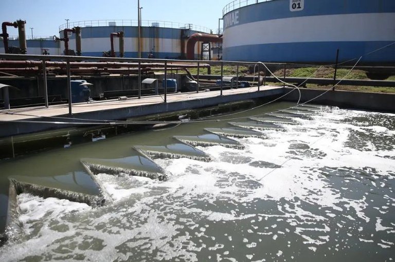 Foto de uma estação de tratamento de água ou esgoto. Em primeiro plano, um tanque aberto contém água turva e espumosa, com uma série de estruturas triangulares (vertedouros) por onde a água flui, criando agitação. Acima deste tanque, há uma passarela de concreto com guarda-corpos e alguns equipamentos ou motores, além de tubulações vermelhas. Ao fundo, destacam-se grandes tanques de armazenamento cilíndricos, pintados de azul e branco. O céu está azul e claro.