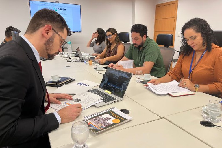 foto de grupo de profissionais em uma reunião de trabalho, sentados ao redor de uma mesa de conferência com documentos e laptops.