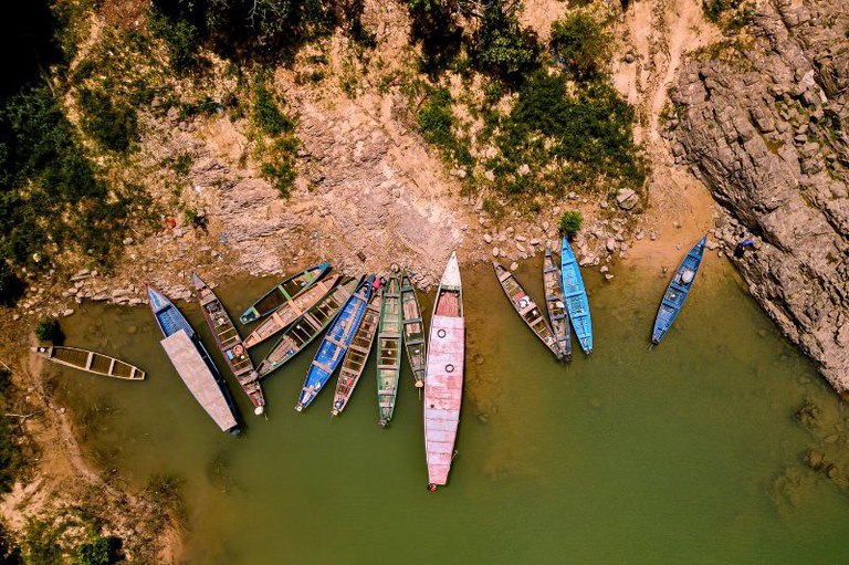 Esta fotografia aérea apresenta uma vista zenital de diversos barcos estreitos e coloridos atracados na margem de um rio de águas esverdeadas, na região do Pedral do Lourenção. As embarcações, que variam em tons de azul, verde, rosa e madeira natural, estão dispostas de forma irregular ao longo de uma borda rochosa e seca, criando um contraste visual vibrante com a textura árida do solo e a vegetação esparsa no topo da imagem. A composição destaca a calmaria das águas e a organização orgânica dos barcos, sugerindo uma pausa na rotina ribeirinha em um cenário marcado por formações rochosas características.