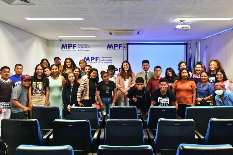 A foto mostra um grupo diverso de pessoas reunidas em uma sala de conferências com cadeiras azuis e pretas. No fundo, há um painel branco com o logotipo do MPF (Ministério Público Federal) e o texto "Procuradoria da República em Santarém". Uma tela de projeção azul está ao lado do painel, com bandeiras posicionadas ao fundo. O grupo inclui homens e mulheres de diferentes idades e etnias, alguns sorrindo, outros com expressões neutras. Algumas pessoas vestem roupas casuais, enquanto outras estão mais formais, incluindo um homem e uma mulher com terno.