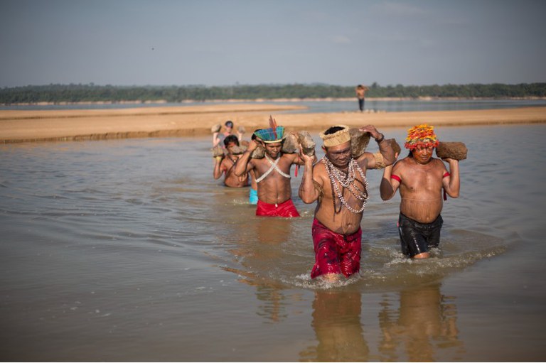 A imagem mostra um grupo de pessoas indígenas caminhando em uma área de água rasa do rio Tapajós, carregando grandes pedras nos ombros. As pessoas estão usando cocares, colares, e peças de roupa tradicionais. Eles estão de peito nu e vestem calças curtas. Ao fundo, mais pessoas seguem na mesma linha. A paisagem ao redor é aberta, com uma área de praia arenosa visível ao fundo e uma faixa de floresta distante no horizonte. O céu está claro e ensolarado.