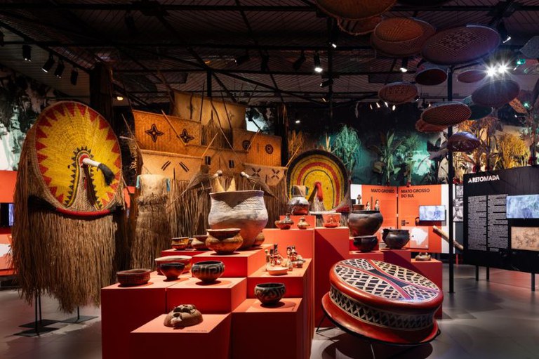 Foto de exposição em museu com artefatos indígenas, incluindo cerâmicas em bases vermelhas, um cocar grande, e cestaria pendurada no teto.