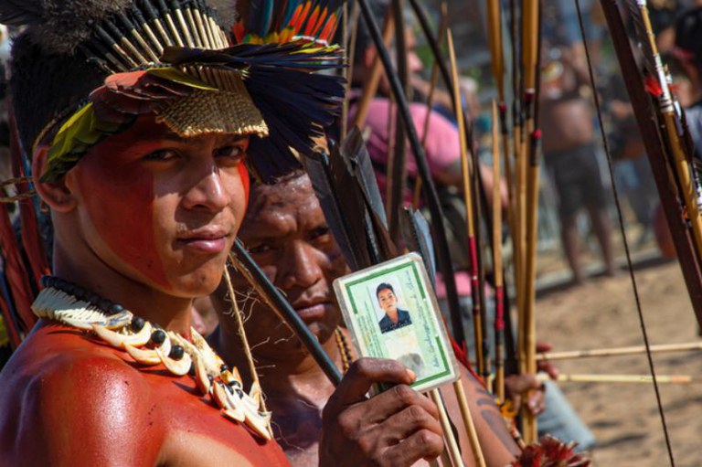 Um jovem indígena com o rosto e o peito pintados de vermelho, usando um cocar de penas e um colar, olha para a câmera enquanto segura um documento de identidade brasileiro (RG). Ao fundo, outros indígenas estão reunidos, muitos com arcos e flechas