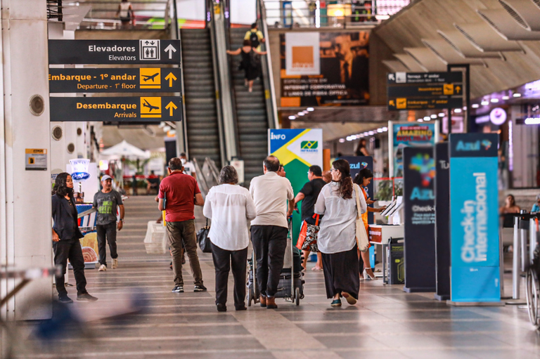 Foto do saguão do aeroporto com pessoas caminhando. Placas indicam Elevadores, Embarque - 1º andar e Desembarque. Há escadas rolantes e totens de empresas aéreas ao fundo em um ambiente amplo e claro.