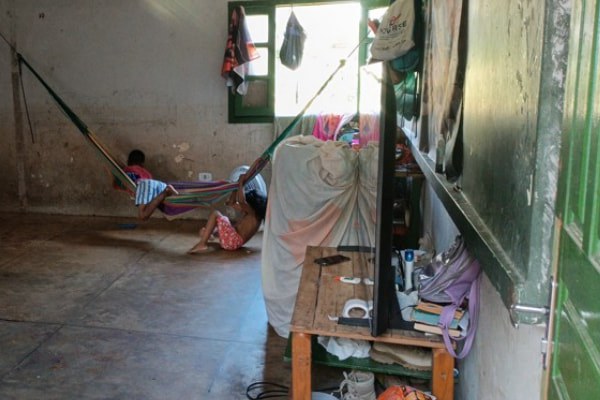 Foto retangular de cômodo interno simples com duas crianças: uma em rede listrada, outra no chão. Ao fundo, janela com esquadria verde; à direita, mesa com objetos, incluindo uma TV