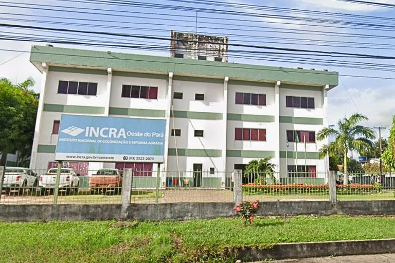 Fachada do prédio do INCRA Oeste do Pará, um edifício de três andares branco e verde, com uma grande placa de identificação azul na frente, visto da rua