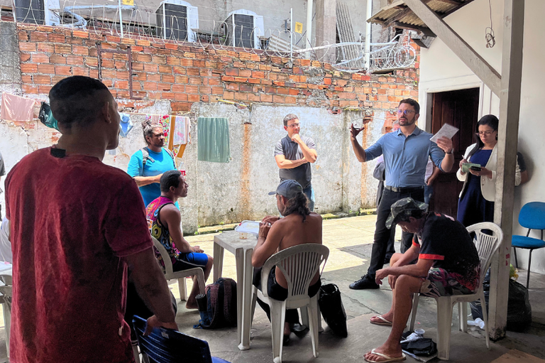 A foto mostra um grupo de pessoas reunidas em um pátio externo com paredes de tijolos aparentes e arame farpado no topo. O procurador da República está de camisa azul, em pé, gesticulando e falando com outros homens sentados ou em pé.