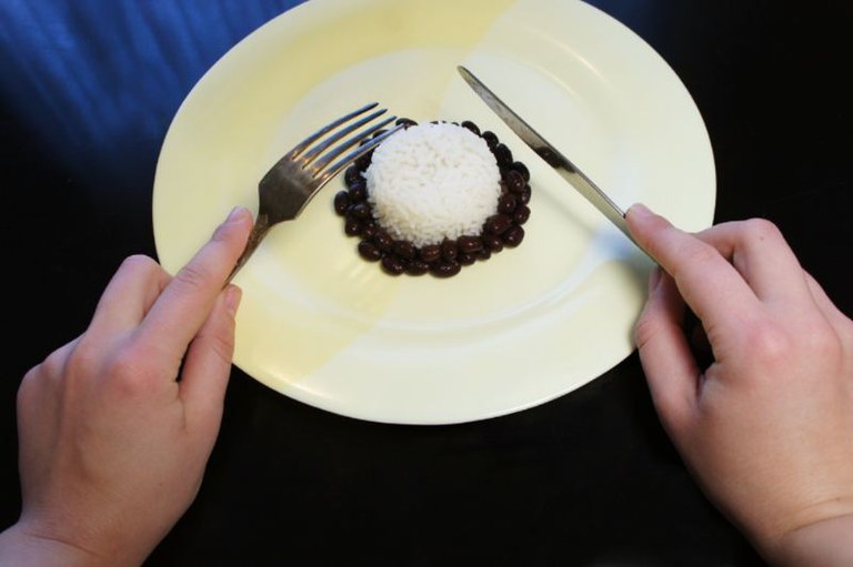 Foto de mãos que seguram garfo e faca em um prato com pequena porção de feijão preto e uma bola de arroz no centro.
