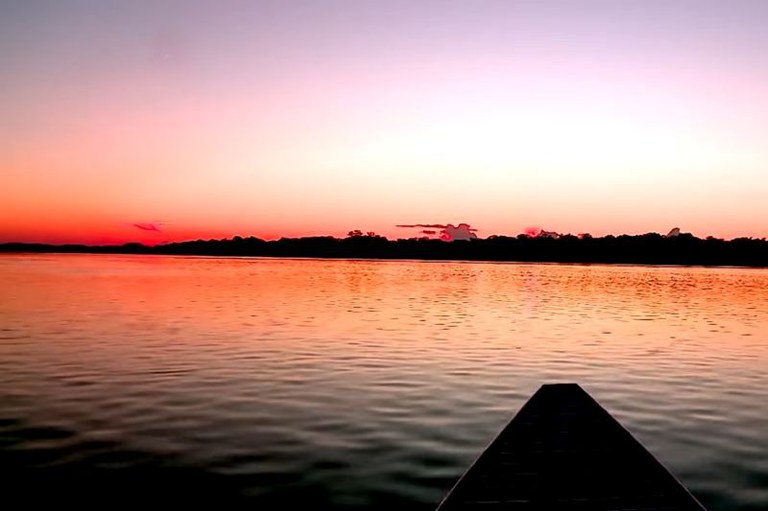 Foto de um pôr do sol sobre um rio, com o céu tingido de tons de laranja e rosa. No horizonte, há uma linha com silhuetas de árvores das margens do rio. A ponta de um barco é visível no canto inferior direito da imagem, apontando em direção ao horizonte.