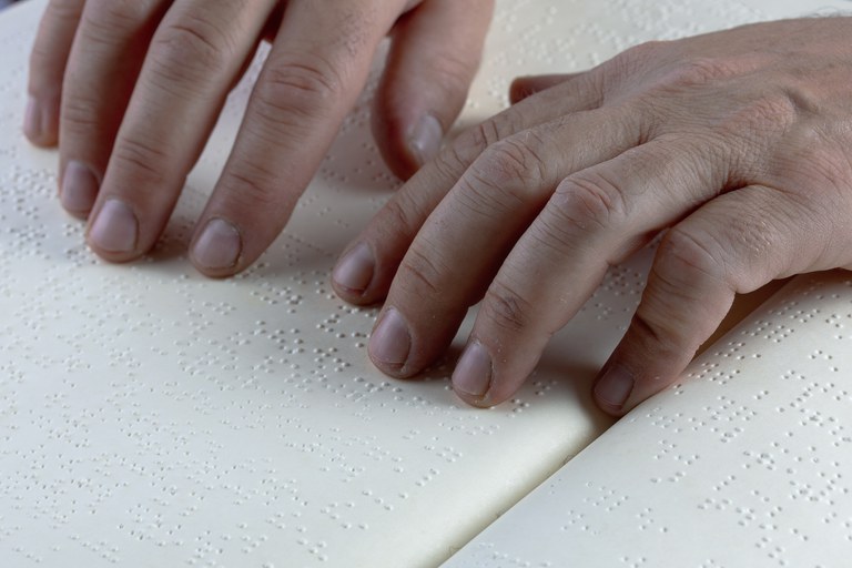 A imagem mostra um close de duas mãos com os dedos posicionados sobre as páginas de um livro aberto. As páginas são brancas e estão repletas de pequenos pontos em relevo, característicos do sistema de leitura Braille.