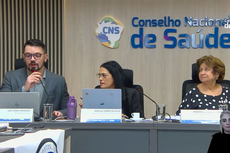 captura de tela mostra três pessoas sentadas em bancada sob logo do Conselho Nacional de Saúde. Sadi Flores Machado fala ao microfone. Priscila Torres da Silva e Francisca Silva observam. Intérprete de Libras no canto