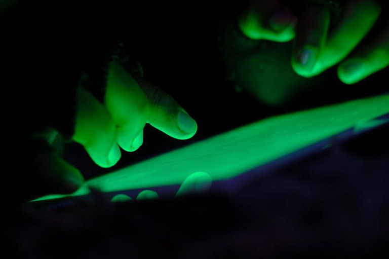 Foto que mostra dedos de mãos interagindo com uma superfície iluminada por uma luz verde intensa, de tela de dispositivo eletrônico. O fundo está escuro, destacando a luminosidade verde.