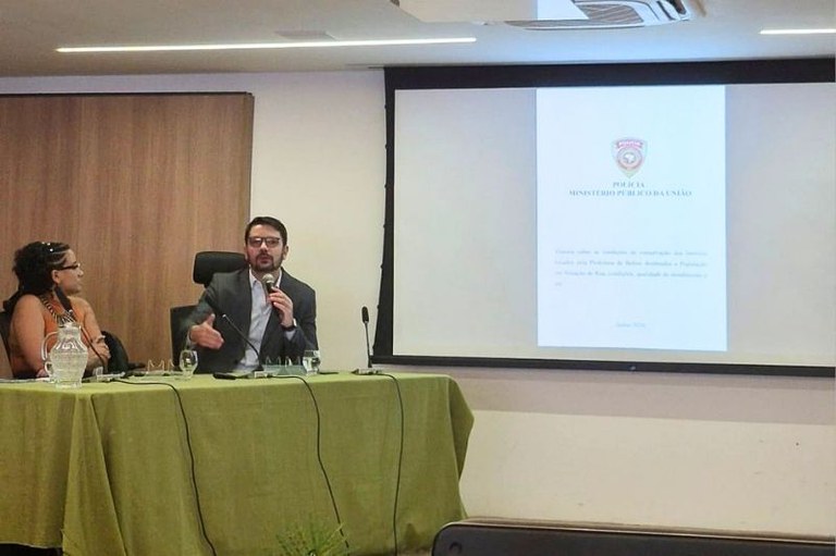 Homem de terno fala ao microfone sentado à mesa verde com uma mulher ao lado. Ao fundo, tela projeta um documento oficial.