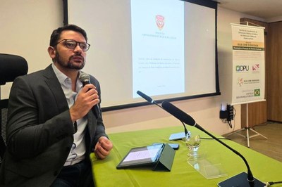  Homem de óculos e terno segura microfone diante de mesa verde com tablet, microfones e copo. Ao fundo, uma tela projetada e um banner.