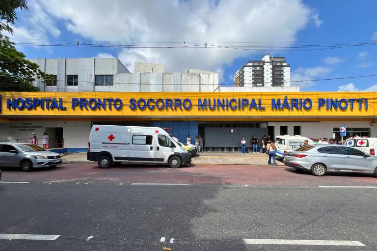 Fachada do Hospital Pronto Socorro Municipal Mário Pinotti com letreiro azul em fundo amarelo, ambulância e carros estacionados na frente.