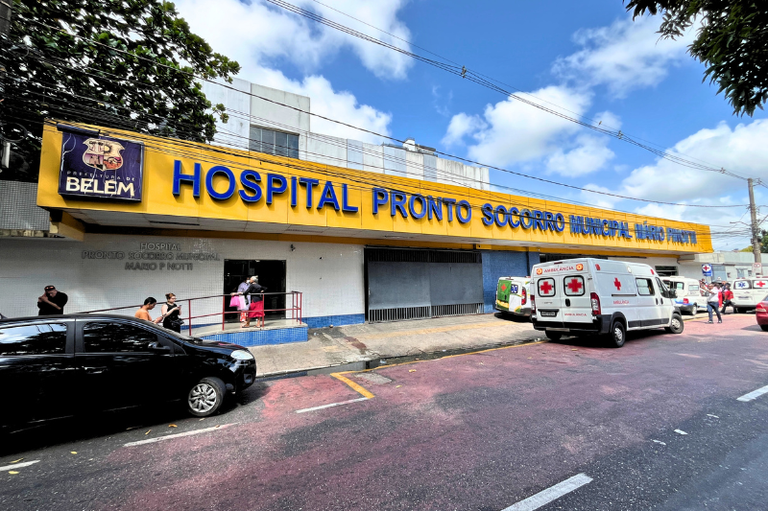 Fachada do Hospital Pronto Socorro Municipal Mário Pinotti, em Belém, com letreiro azul em fundo amarelo. Há ambulâncias brancas estacionadas e pessoas na calçada sob um céu com nuvens claras