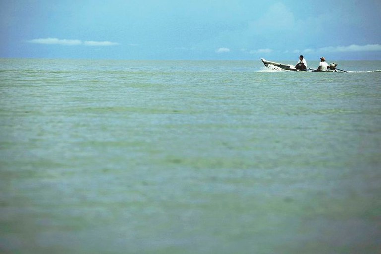 Foto do Rio Tapajós, vasto e esverdeado sob um céu azul. No horizonte, à direita, um pequeno barco a motor com dois homens a bordo atravessa a água.
