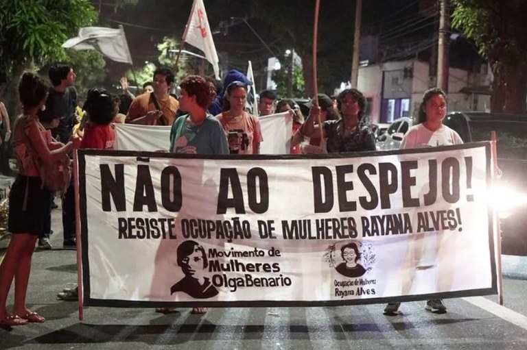 Pessoas seguram em uma rua à noite uma faixa com a frase "Não ao despejo! Resiste Ocupação de Mulheres Rayana Alves!"