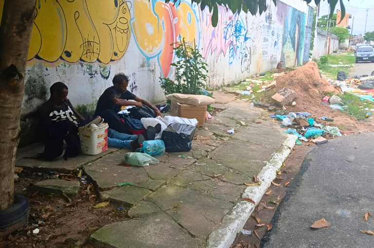 Foto mostra duas pessoas sentadas na calçada, ao lado de um muro com grafites coloridos. Há sacolas plásticas, roupas e objetos espalhados ao redor delas. À direita, há lixo acumulado e entulho próximo à rua, que tem um carro estacionado ao fundo. Há árvores e vegetação ao redor.