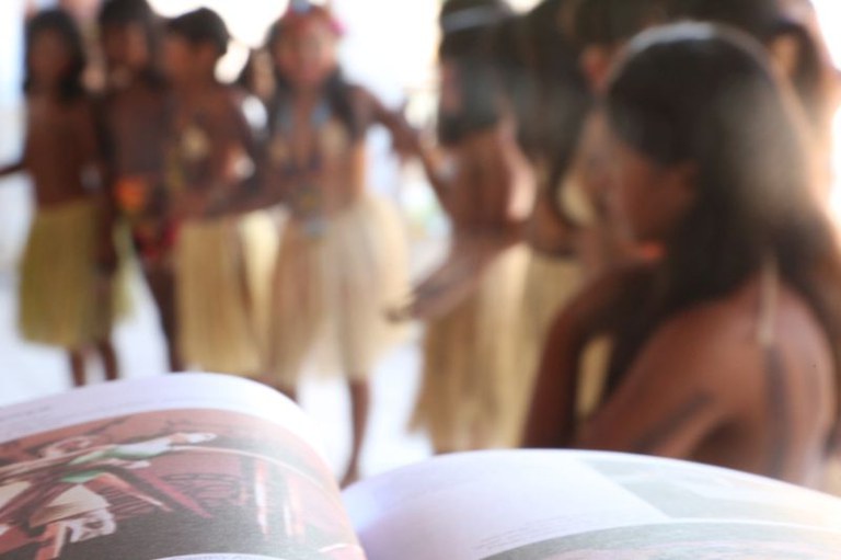 Foto mostra crianças indígenas desfocadas e, em primeiro plano, um livro escolar; no centro, há a expressão Educação escolar indígena