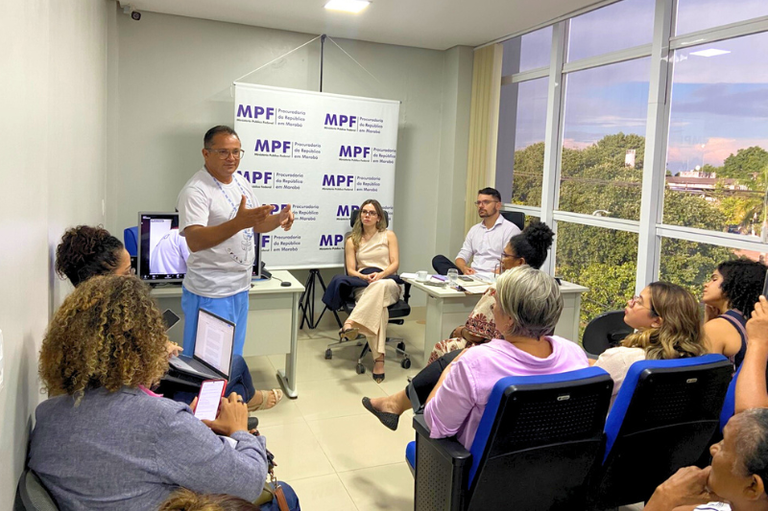 Foto de reunião de pessoas em sala em que, ao fundo, há banner com a logo do MPF. As pessoas estão voltadas para uma pessoa que fala em pé. A sala tem janela com vista para copas de árvores e telhados de casas.