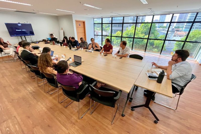 foto de sala de reuniões com 16 pessoas sentadas em volta de uma longa mesa de madeira.