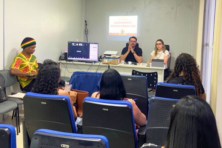 Foto de uma reunião com várias pessoas sentadas em cadeiras azuis, duas pessoas à mesa, e projeção em tela branca.