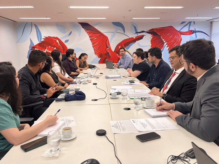 #ParaTodosVerem: Foto de reunião em mesa branca com várias pessoas sentadas. Ao fundo, painel com guarás vermelhos. Sobre a mesa, papéis, copos d'água, xícaras de café, notebooks e celulares.