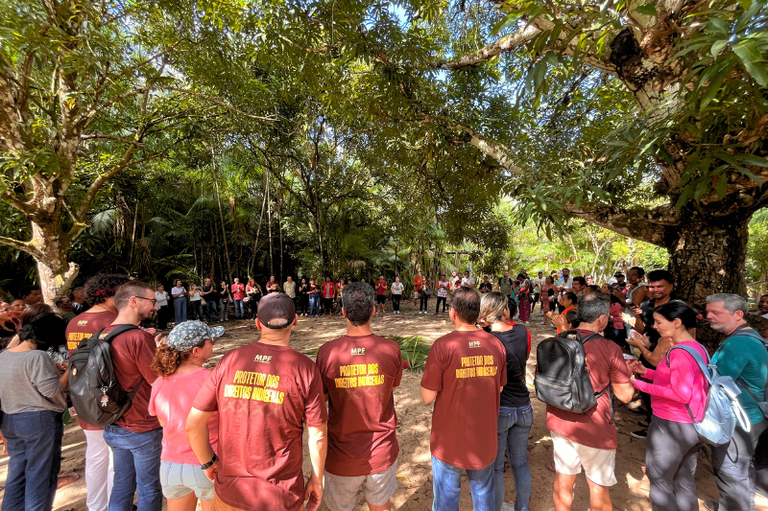 #ParaTodosVerem: Em uma área aberta cercada por árvores, várias pessoas estão reunidas em círculo, muitas vestindo camisetas bordô com os dizeres "MPF Protetor dos Direitos Indígenas" nas costas. Algumas pessoas usam mochilas e roupas casuais, e há grande diversidade de posições e conversas. A cena é iluminada pela luz do dia filtrada pelas copas das árvores. Ao fundo, há mais pessoas compondo o círculo sob a vegetação densa.