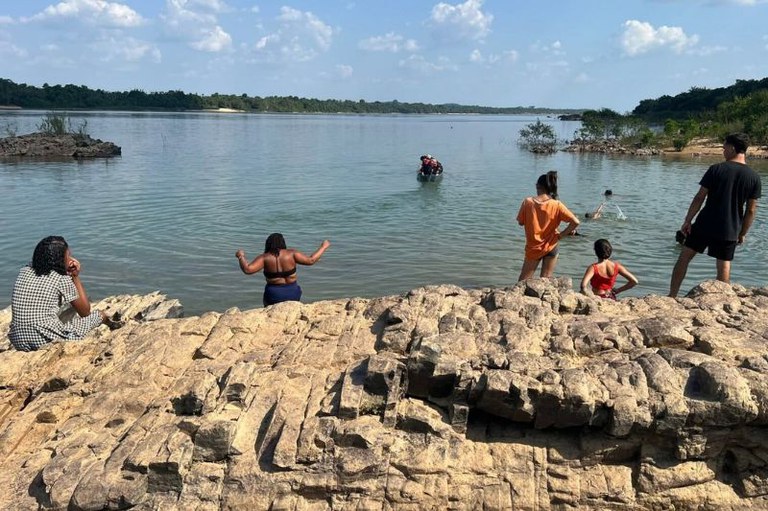 Grupo de pessoas junto à margem de um rio, reunidas numa zona de rochas claras e rugosas num dia ensolarado.

À esquerda, uma pessoa senta-se nas rochas a observar a água, enquanto outra pessoa de fato de banho azul e top preto entra ou prepara-se para mergulhar no rio. À direita, três pessoas estão de pé ou sentadas nas rochas a olhar na direção do rio: uma jovem de t-shirt taranja, outra de fato de banho vermelho e um homem de t-shirt e calções pretos.
Na água, ao fundo, sobressai uma pequena embarcação (canoa ou barco) com pessoas a bordo e algumas pessoas a nadar. A paisagem ao fundo é composta por águas calmas e uma margem repleta de vegetação densa sob um céu azul com nuvens dispersas.