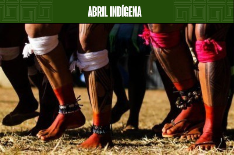 Fotografia ilustrativa mostra, em close (do joelho para baixo), as pernas de indígenas reunidos, evidenciando adereços coloridos e pinturas corporais típicas. Na parte superior da imagem, há uma faixa verde com o texto Abril Indígena.