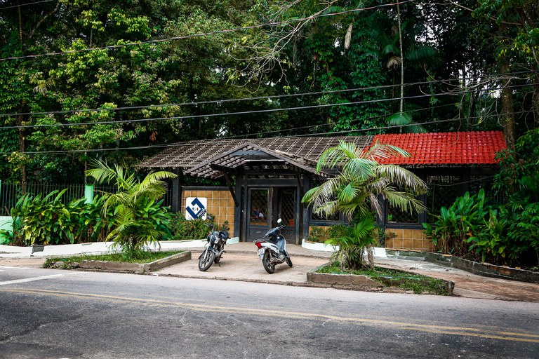 A imagem mostra a entrada de uma das sedes da Fundação Escola Bosque, com estrutura de madeira e telhado inclinado parcialmente coberto por telhas vermelhas. Há um portão de vidro com grade metálica e paredes revestidas de azulejos bege. Duas motos estão estacionadas na frente do local, que é cercado por vegetação densa, incluindo árvores e plantas tropicais. Há uma pequena placa branca e azul fixada na parede à esquerda da entrada. Na calçada, duas palmeiras estão plantadas em canteiros de concreto, e fios elétricos atravessam a imagem no alto.