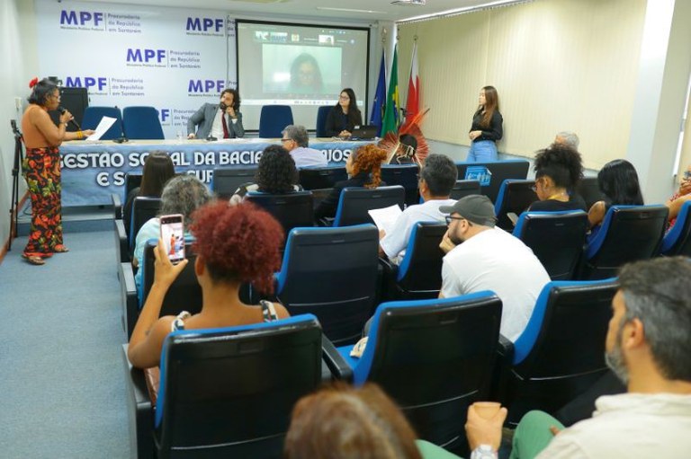 Pessoas sentadas em um auditório assistem a uma apresentação com palestrantes e uma projeção em tela ao fundo.