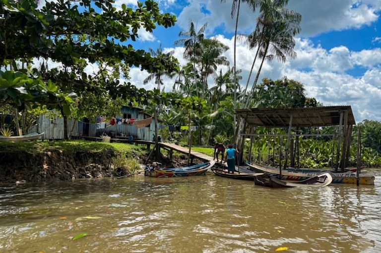 Foto colorida de margem de rio com casa de madeira, palafitas, pontes, barcos pequenos e pessoas. No fundo, palmeiras e céu nublado