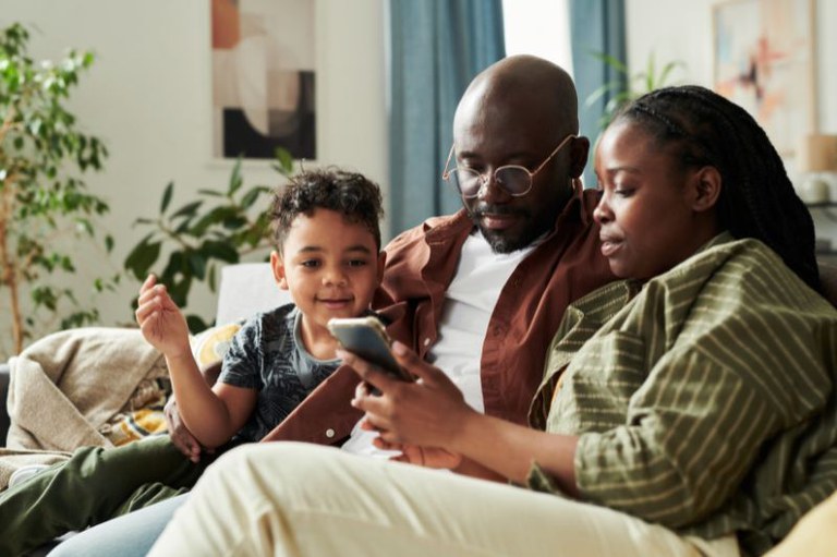 foto de uma família com pai, mãe e filho pequeno, sentada no sofá da sala, olhando para um smartphone.