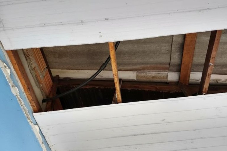 O teto de um cômodo com duas telhas de amianto cinzas expostas por um buraco no forro.