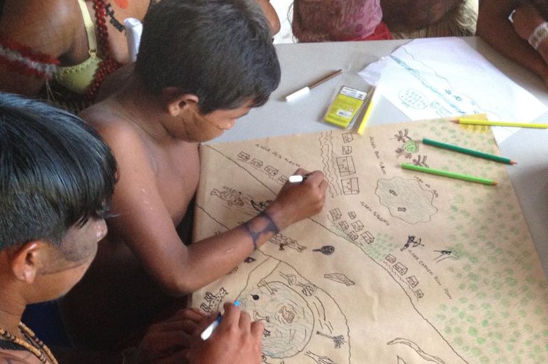 Foto de crianças indígenas, algumas sem camisa e com pinturas corporais, reunidas ao redor de uma mesa desenhando um mapa de seu território em uma grande folha de papel pardo. Elas utilizam canetas e lápis de cor para ilustrar elementos como aldeias, rios, animais e vegetação.