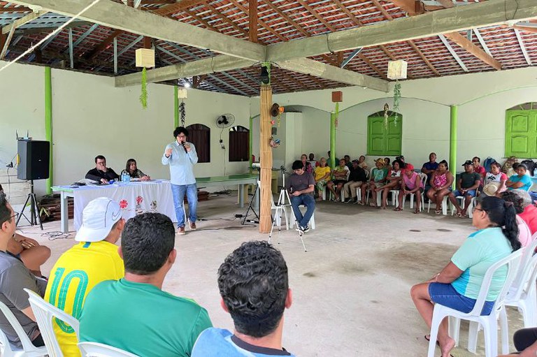Foto em formato retangular. Um homem fala ao microfone para uma plateia sentada em cadeiras brancas, dentro de um salão amplo. Na frente, outras duas pessoas estão sentadas atrás de uma mesa. O ambiente possui teto com telhas aparentes, paredes brancas e detalhes em verde nos pilares e janelas.