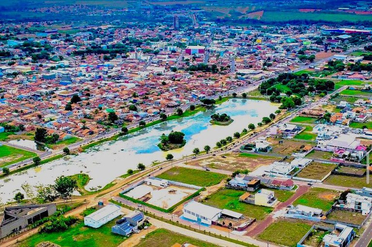 Vista aérea de Paragominas, com lago central contendo duas ilhas arborizadas, cercado por ruas, casas coloridas e vegetação ao fundo.