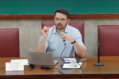 Foto: Cepedisa/USP/Homem branco de óculos, barba e camisa clara fala ao microfone sentado à mesa com notebook e placa de identificação Sadi Machado.