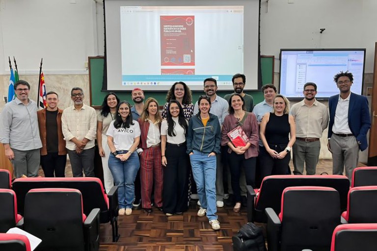  Dezessete pessoas sorriem à frente de um telão projetando a capa de um livro em um auditório com cadeiras pretas e vermelhas.