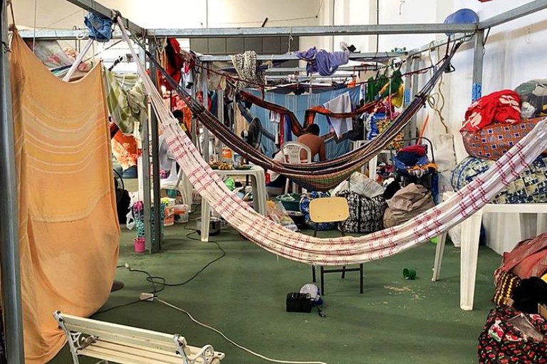 Foto de abrigo improvisado com armação metálica, redes, roupas penduradas e pertences, com uma pessoa sentada ao centro.